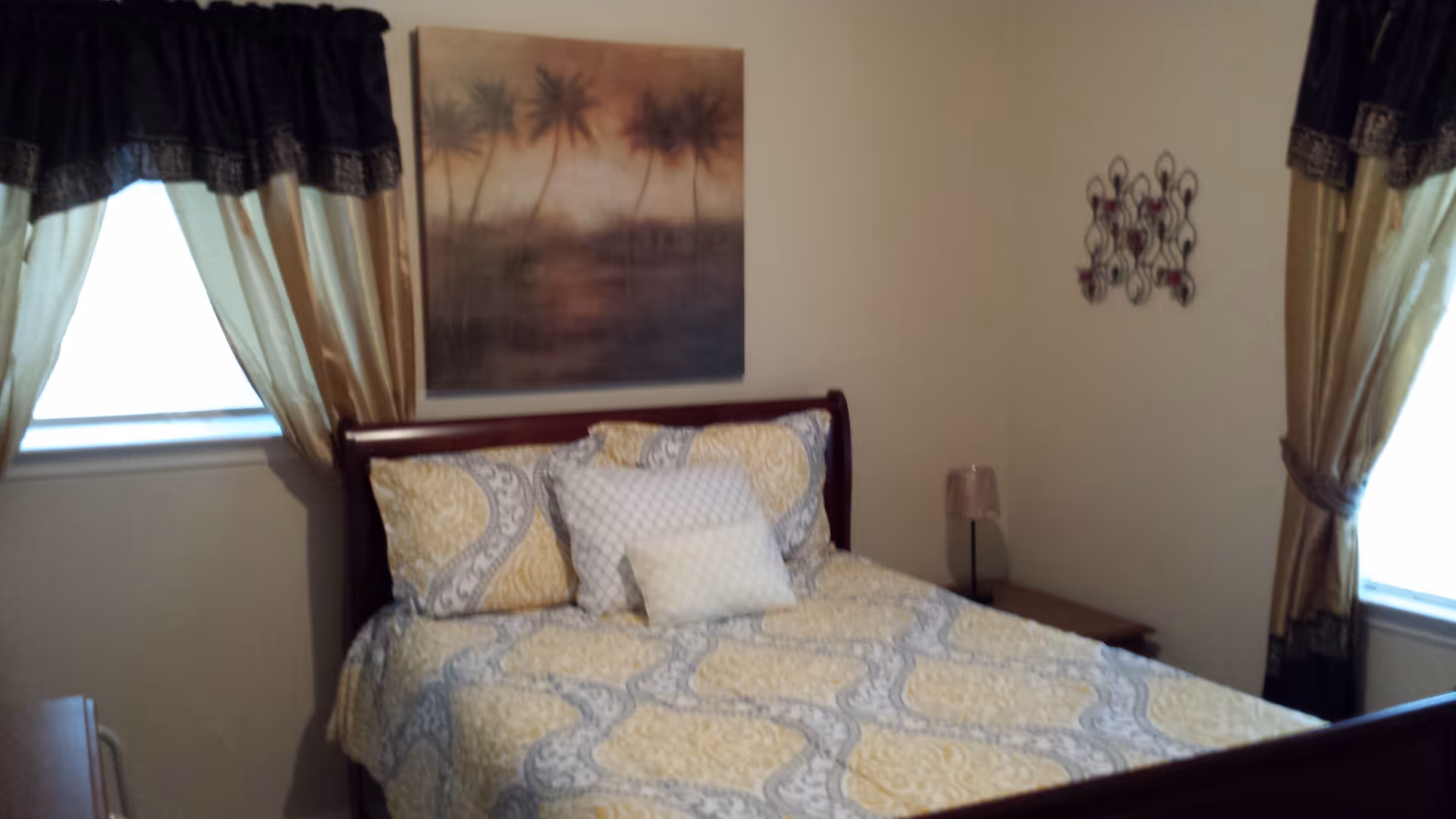 A bedroom with a bed covered in a yellow and gray patterned quilt and several pillows. The room has two windows with gold and black curtains. Above the bed is a painting of palm trees at sunset, and there is a small nightstand with a lamp on the right side of the bed.