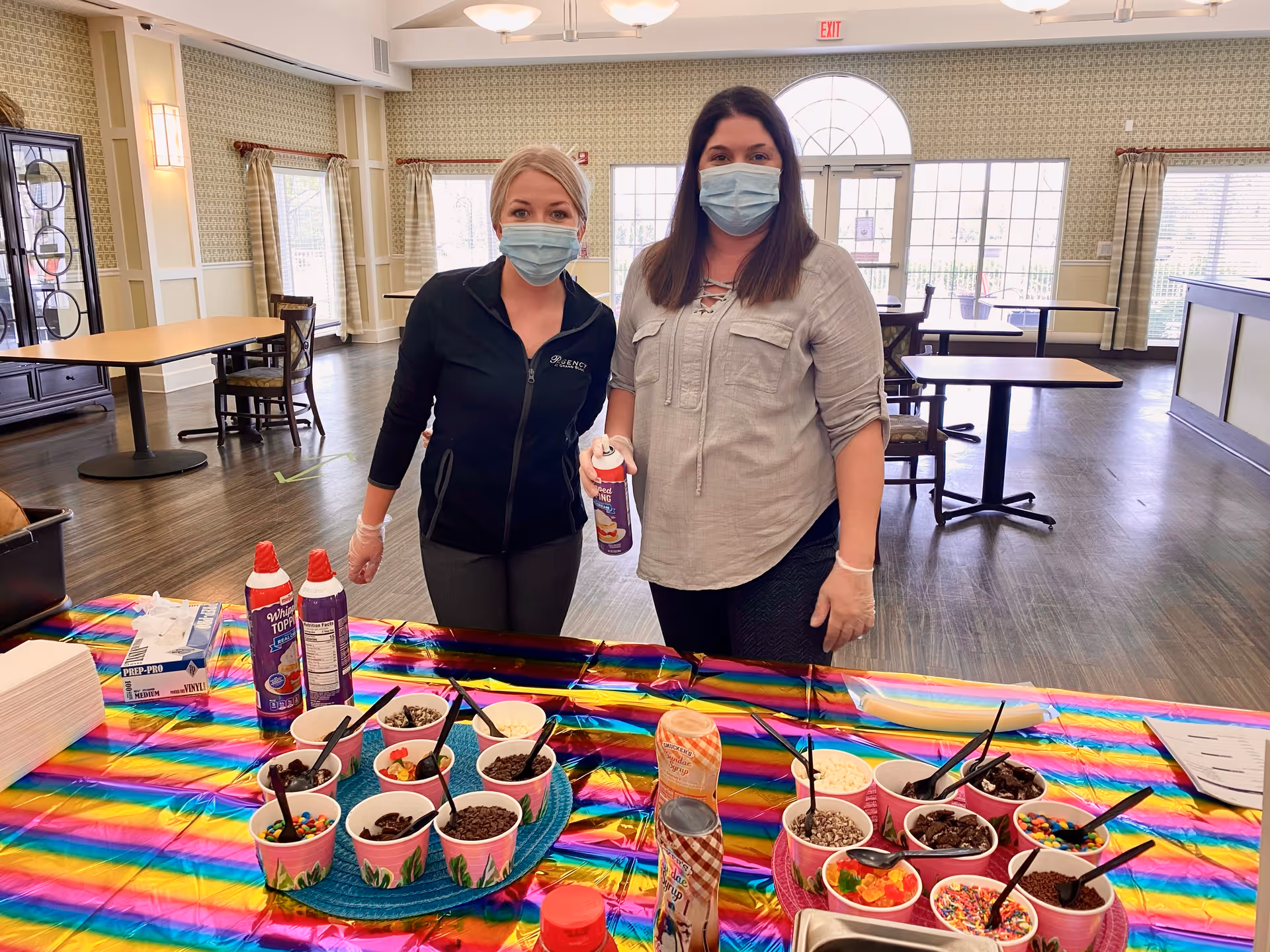 Two women wearing face masks and gloves standing behind a table covered with a colorful striped tablecloth. The table has various cups filled with ice cream toppings such as sprinkles, chocolate chips, and gummy bears, along with cans of whipped topping. The setting appears to be a dining area with tables and chairs, large windows, and patterned wallpaper.