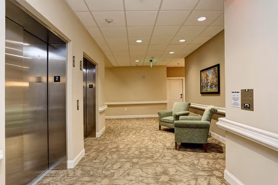 Interior hallway of a senior living facility with two closed stainless steel elevators on the left, beige walls, patterned carpet, two green upholstered armchairs on the right, a framed painting on the wall, and a white door at the end of the hallway.