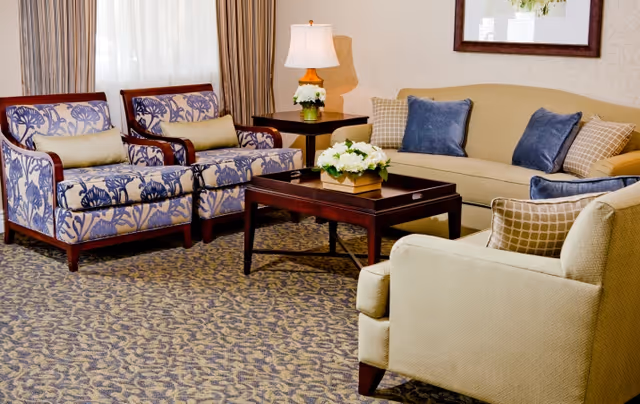 A bright living room with patterned armchairs, a beige sofa, a wooden coffee table topped with a floral arrangement, and a table lamp.