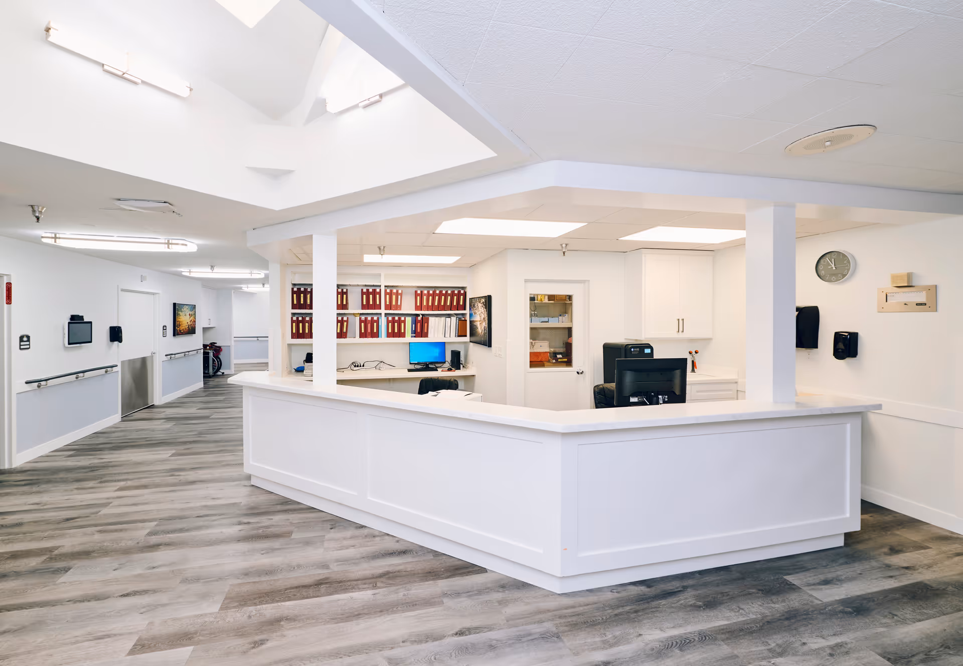 A bright and clean reception desk area inside a senior living facility with white walls, wood-look flooring, and overhead lighting. The desk has computer monitors, office chairs, and shelves with red binders. A hallway extends to the left with handrails along the walls.