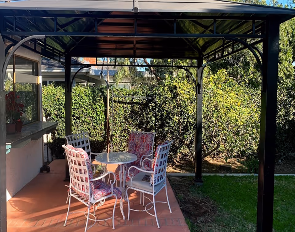 Outdoor covered patio with a metal gazebo, a round glass table and four cushioned metal chairs next to a hedge.