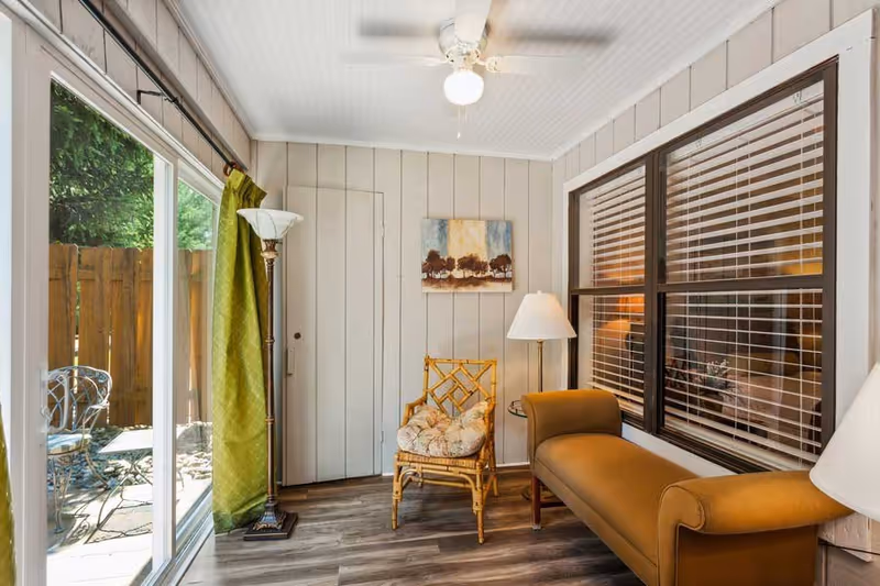 A cozy sunroom with a large sliding glass door leading to an outdoor patio with metal chairs and a table. Inside, there is a cushioned wicker chair, a brown upholstered bench, a floor lamp, and a ceiling fan with a light. The walls are paneled in light wood, and there is a window with blinds looking into another room. A small painting of trees hangs on the wall.