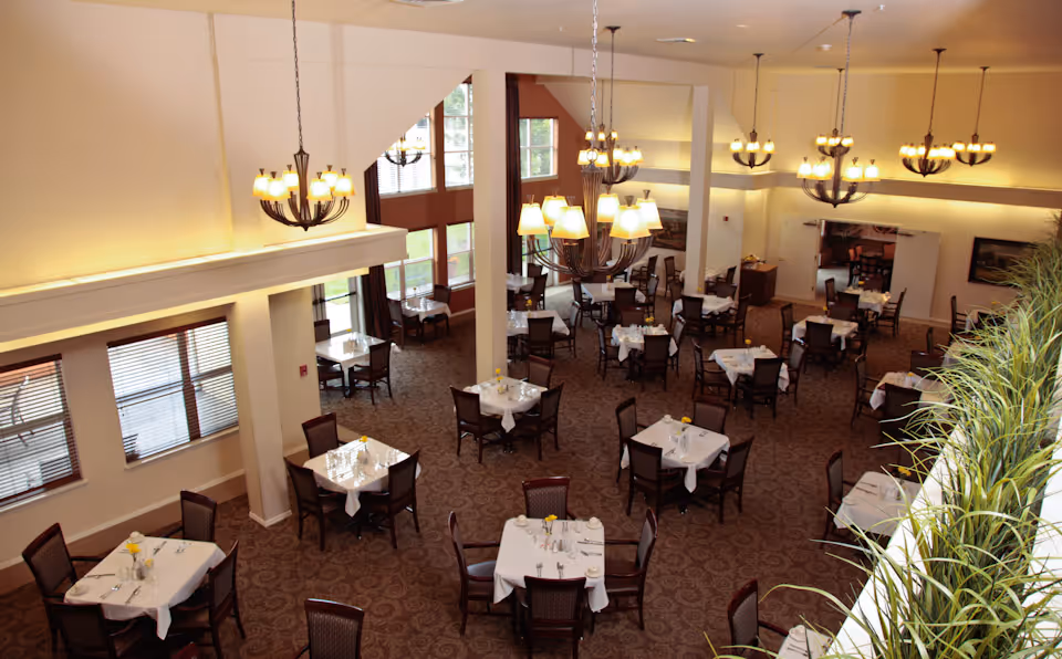 Spacious dining room with multiple tables set with white tablecloths and chandeliers under a high ceiling.