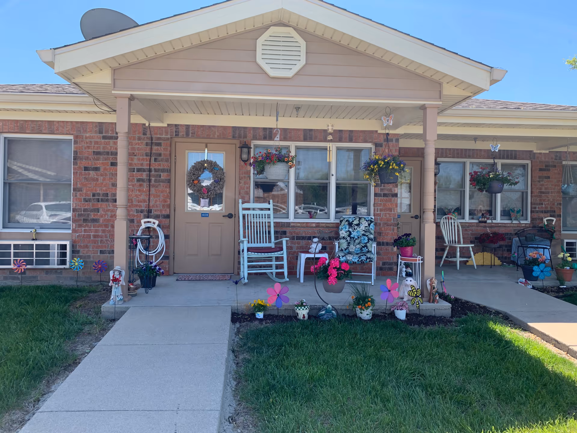 Front entrance of a brick single-story building with a covered porch, rocking chairs, hanging baskets and potted flowers along a concrete walkway.