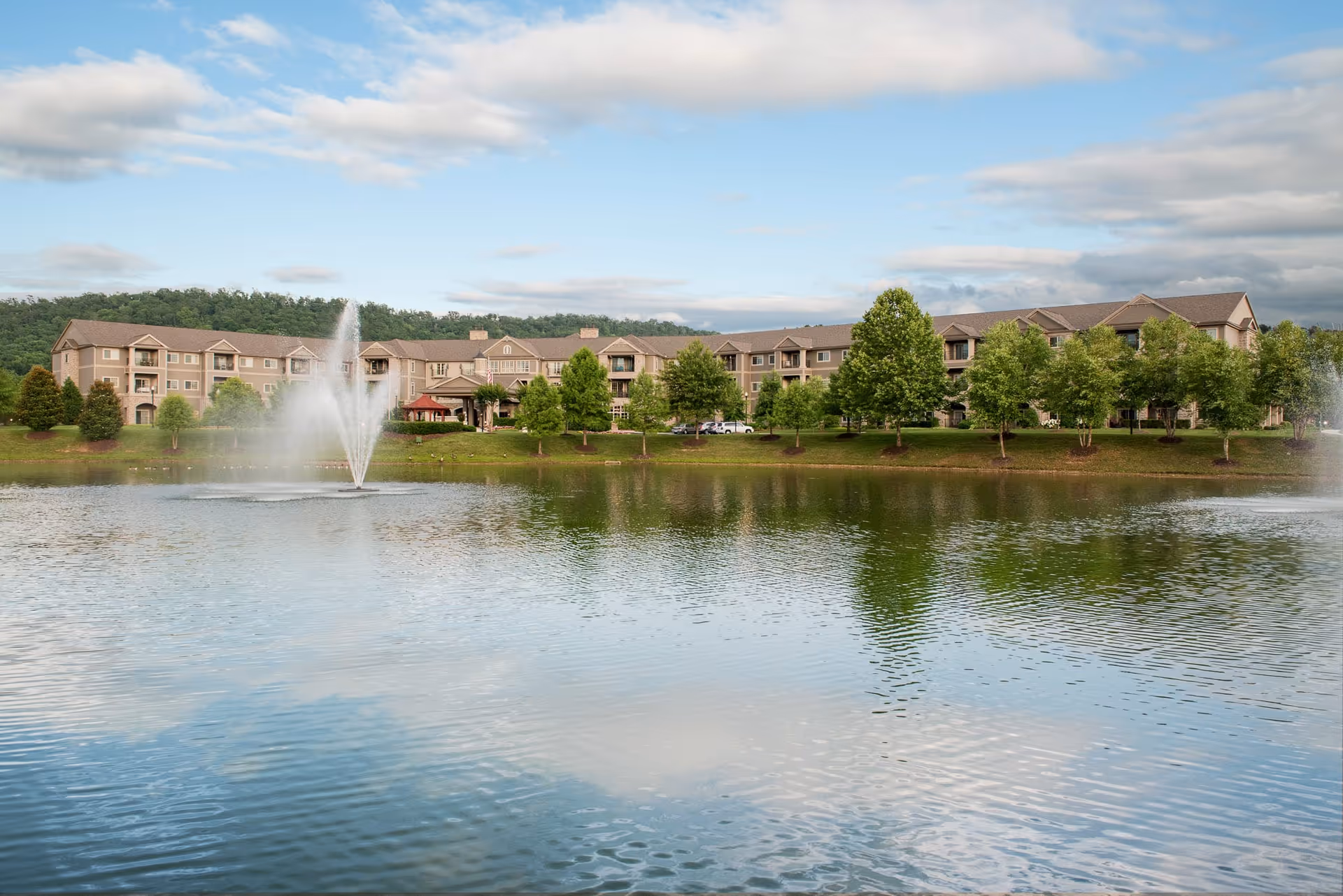 A large senior living facility building named Garden Plaza Of Greenbriar Cove is visible across a pond with two water fountains. The building is surrounded by green trees and a grassy area under a partly cloudy sky.