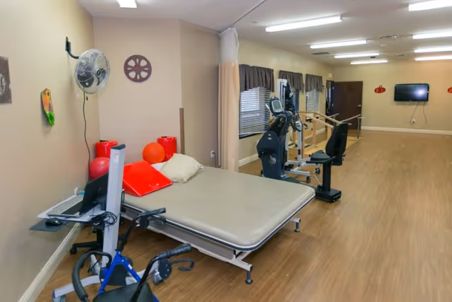 A rehabilitation room with a therapy bed, exercise equipment including a stationary bike and parallel bars, a wall-mounted fan, and a wall-mounted TV. The room has wood flooring, beige walls, and windows with dark curtains.