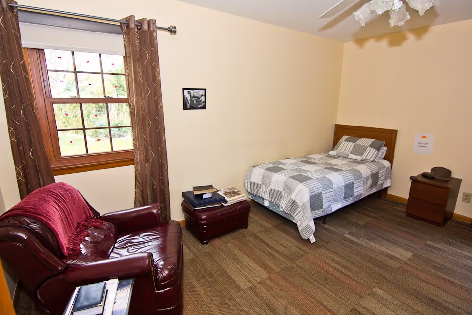 A simple bedroom with a single bed covered in a gray and white checkered quilt, a wooden headboard, and a matching pillow. Next to the bed is a small wooden nightstand with a hat and a remote control on it. Across from the bed is a dark red leather armchair with a red throw draped over the back. A small ottoman in front of the armchair holds a few books and magazines. The room has a window with brown curtains and a beige wall with a small black and white framed photo. The floor is carpeted with a multi-tone brown pattern.