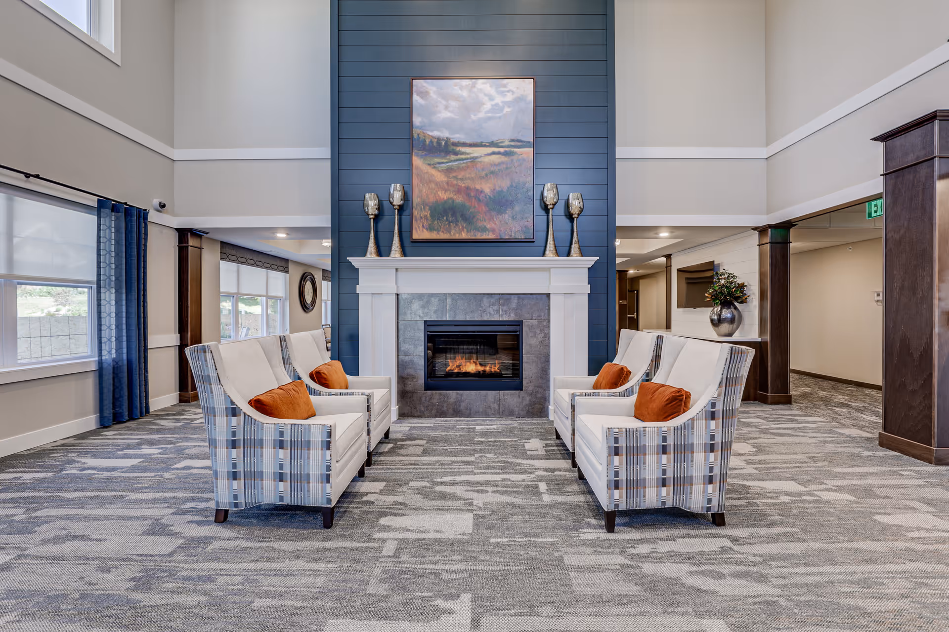 A spacious and well-lit living room area with two sets of plaid armchairs facing each other across a carpeted floor. The room features a modern fireplace with a white mantel and a blue shiplap accent wall above it, decorated with a landscape painting and four decorative candle holders. Large windows with blue curtains allow natural light to fill the space.