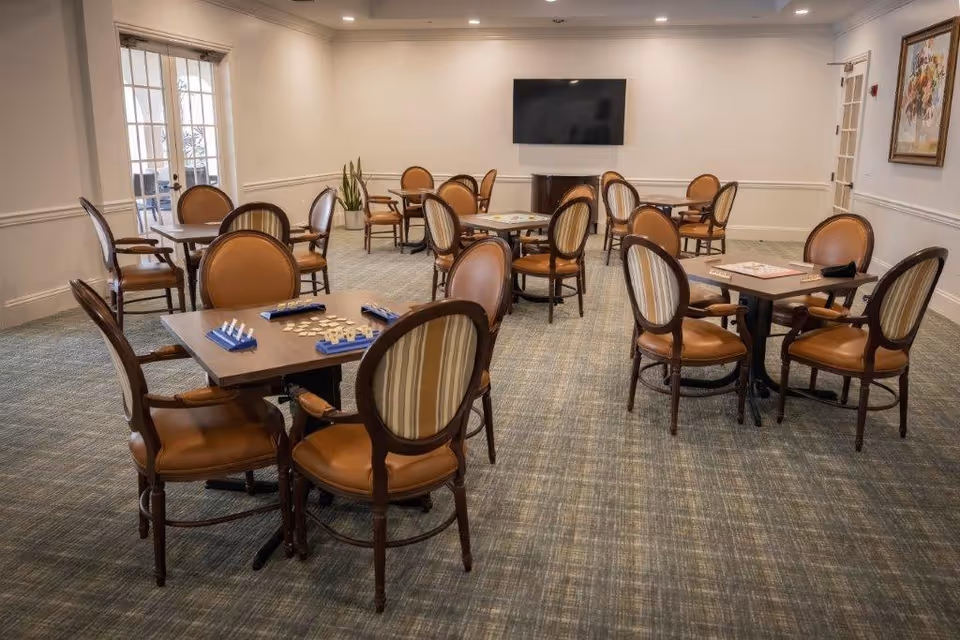 A well-lit room with multiple small square tables, each surrounded by four cushioned chairs with wooden frames. Some tables have game pieces on them, suggesting a recreational or game room setting. The room has carpeted flooring, light-colored walls with molding, a flat-screen TV mounted on the far wall, and a framed painting on the right wall. French doors on the left side lead to an outdoor area.