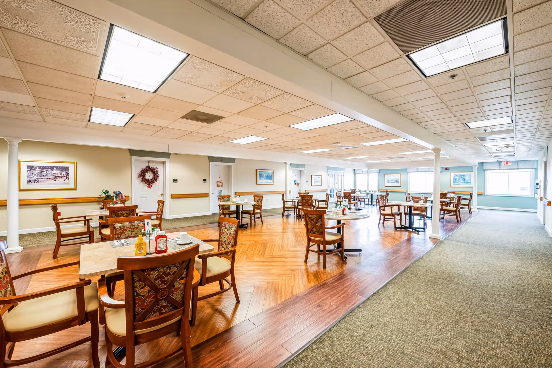 A bright and spacious dining room in a senior living facility with multiple wooden tables and chairs arranged neatly. The room features a combination of carpet and wood flooring, ceiling lights, framed artwork on the walls, and large windows allowing natural light to enter.