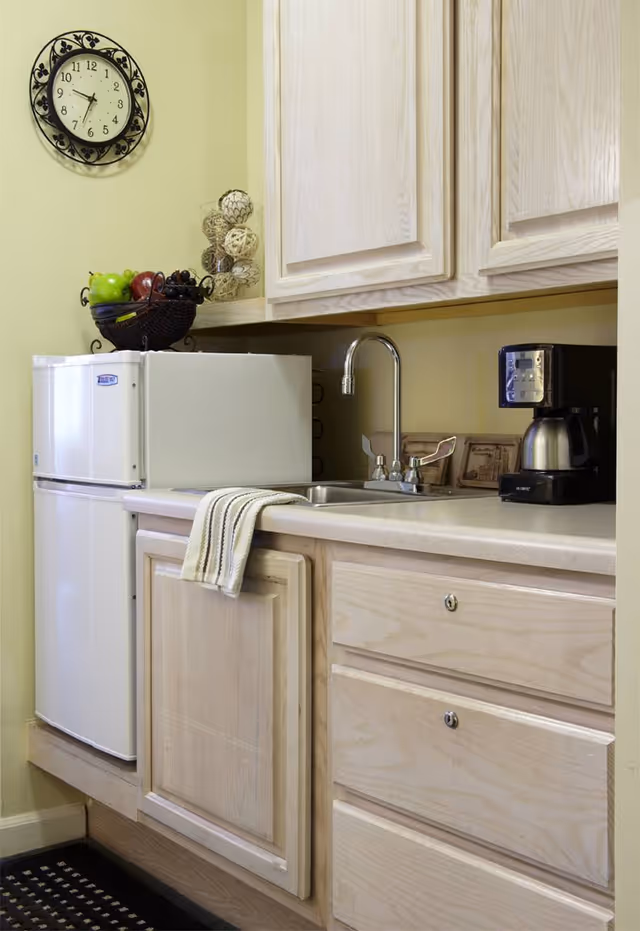 Small kitchen area with light wood cabinets, a white mini refrigerator, a stainless steel sink with a faucet, a coffee maker, a wall clock, and a fruit basket on the counter.