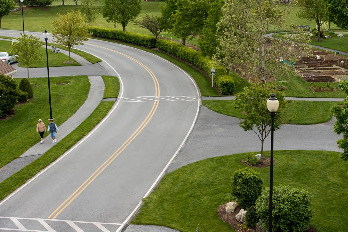A curved road with sidewalks on both sides, surrounded by well-maintained green lawns, trees, and bushes. Two people are walking on the sidewalk on the left side of the road. There are street lamps and a speed limit sign indicating 20 mph. In the background, there are garden plots and more greenery.