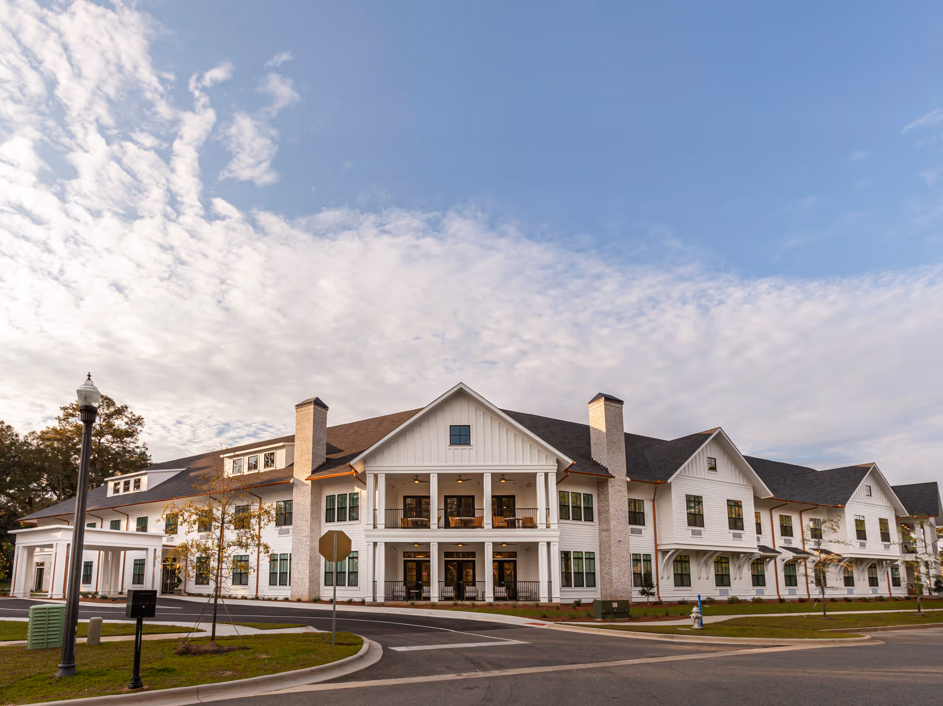Exterior view of a large, white, two-story assisted living and memory care facility building with multiple windows, two chimneys, and a covered entrance. The sky is partly cloudy with blue patches visible.