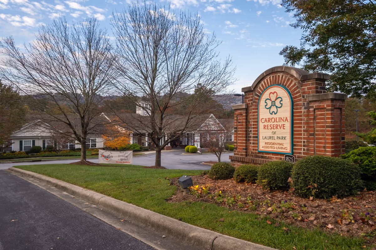 Exterior view of Carolina Reserve of Laurel Park residential assisted living facility with a brick sign in the foreground and the building in the background surrounded by trees and landscaping under a partly cloudy sky.
