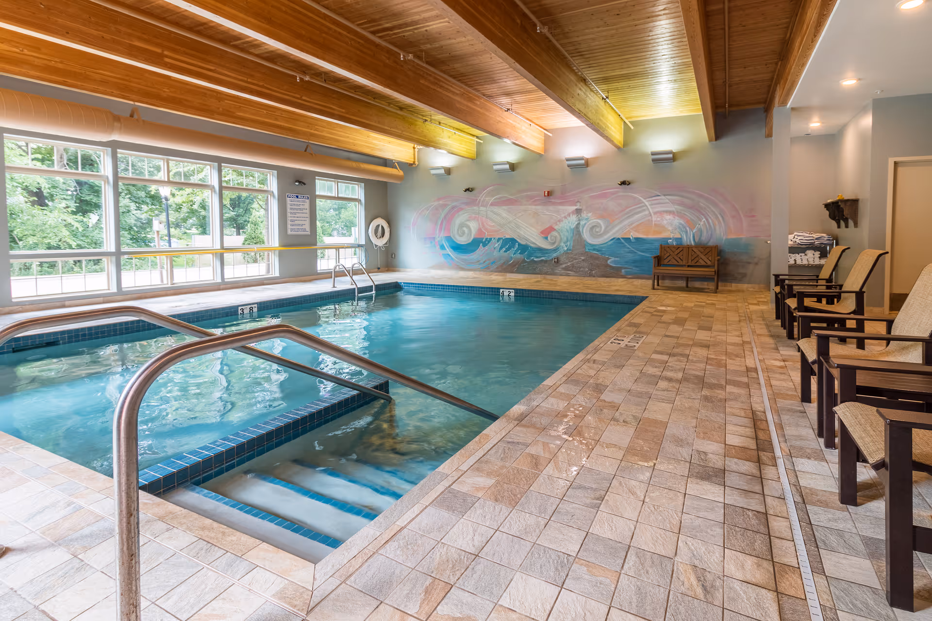Indoor swimming pool with tiled deck, metal handrails, lounge chairs and a colorful mural on the far wall.