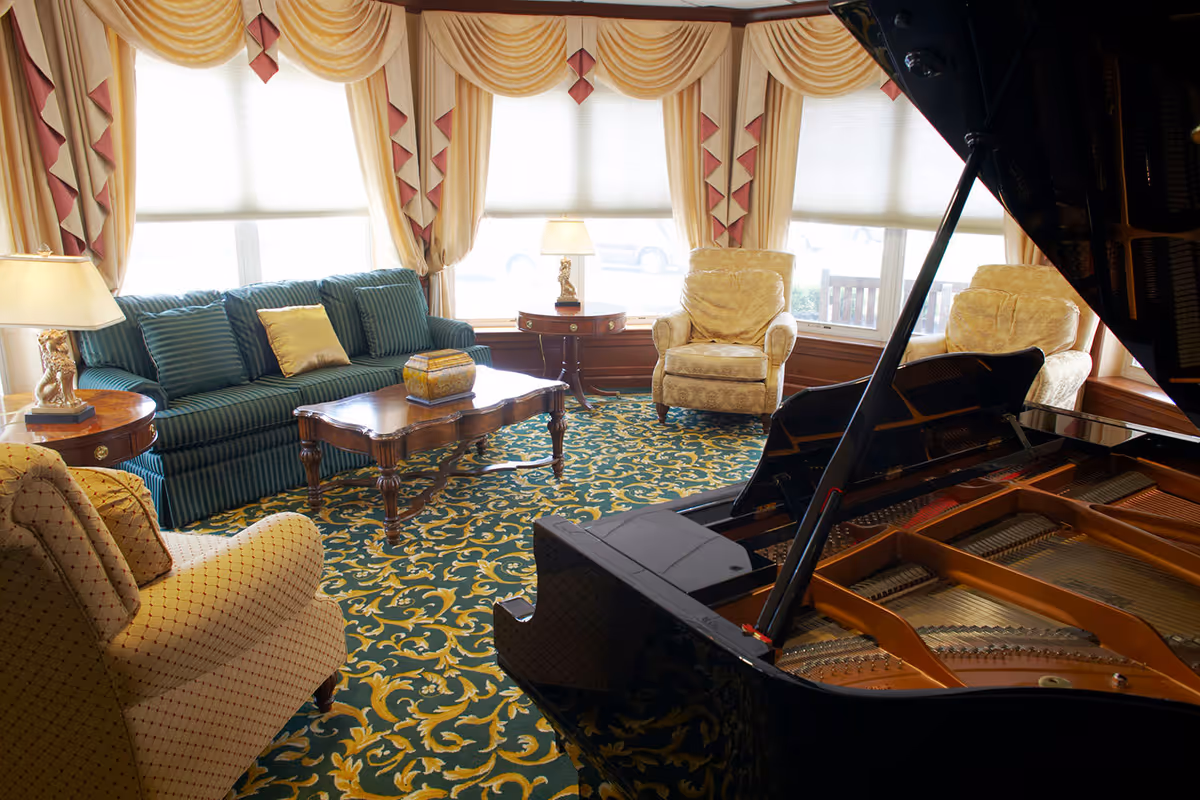 A cozy living room with a green striped sofa, three beige armchairs, a wooden coffee table, two side tables with lamps, large windows with yellow and red drapes, and a grand piano with its lid open in the foreground.