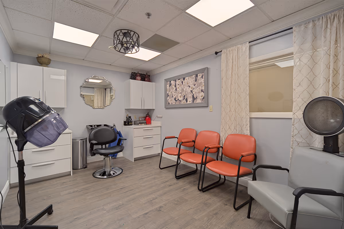 Interior view of a small salon or hair care room with a hair dryer chair, a black salon chair, white cabinets, a decorative mirror, and four chairs with orange and gray upholstery along the wall. The room has light gray walls, wood flooring, a ceiling light fixture, and a window with patterned curtains.