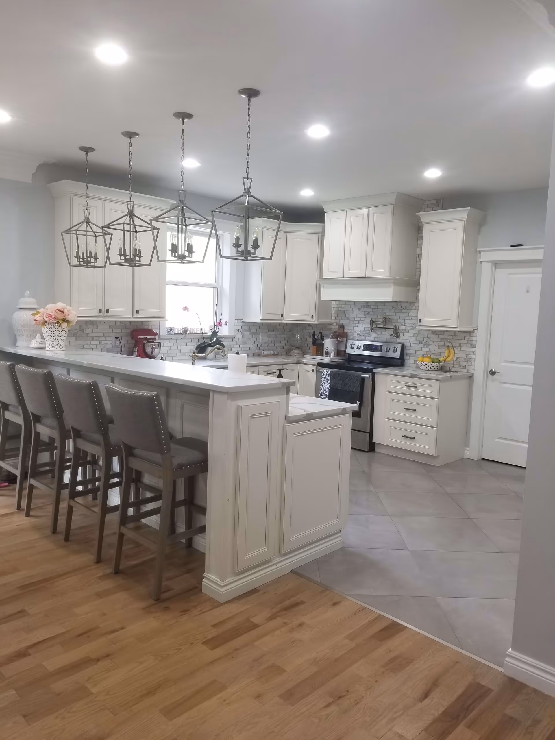A modern kitchen with white cabinetry and a white countertop island with four gray cushioned bar stools. The kitchen features stainless steel appliances, including an oven and stove, and a white tile backsplash with a brick pattern. Four geometric pendant lights hang above the island. The floor transitions from light wood in the foreground to large gray tiles in the kitchen area. There is a window above the sink letting in natural light, and decorative items such as flowers and fruit bowls are placed on the counters.