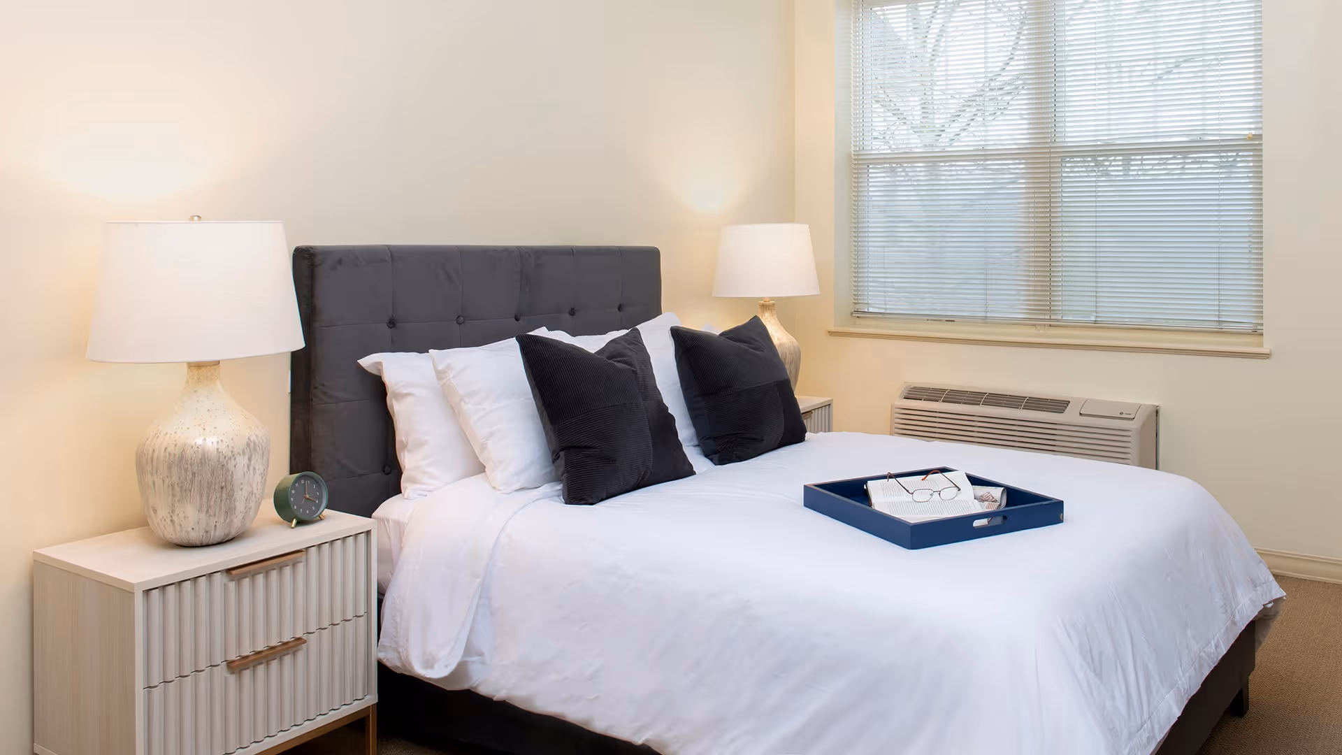 A neatly made bed with white linens and four pillows, two white and two black, in a bedroom. There is a blue tray on the bed holding a pair of glasses and a book. On either side of the bed are matching nightstands with large table lamps. A window with blinds and an air conditioning unit is visible on the right wall.