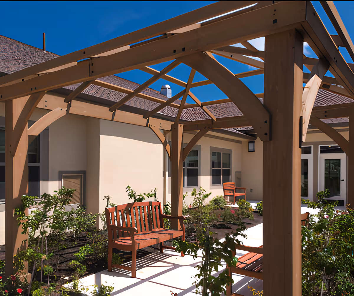 Outdoor courtyard area with wooden pergola structure, red wooden benches, and landscaped garden beds with green plants and flowers, adjacent to a beige building under a clear blue sky.