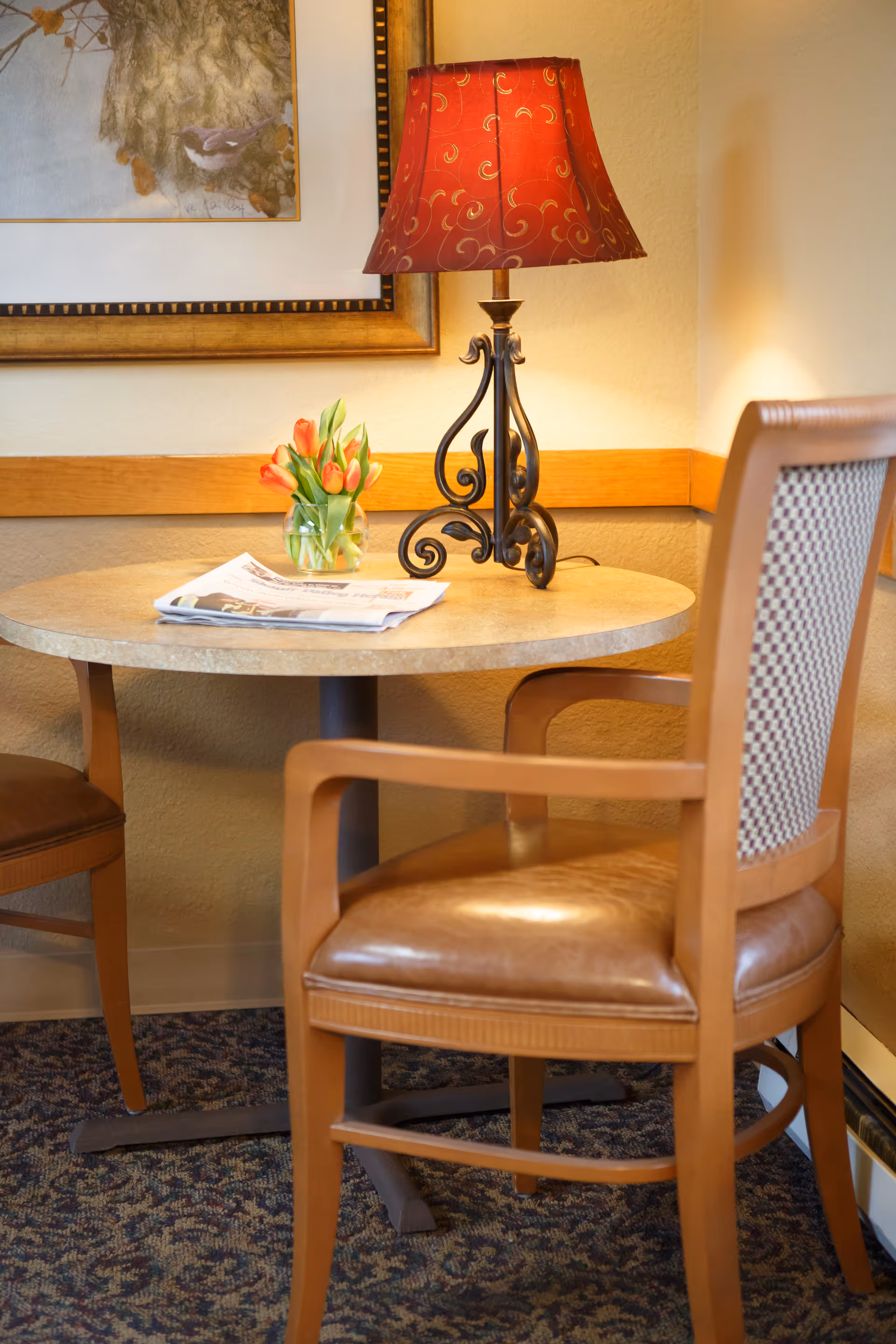 Small round table in a cozy interior corner with a decorative lamp, vase of tulips, a newspaper and two chairs.