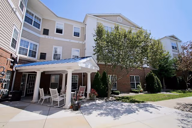 Exterior view of a senior living facility building with a covered patio area featuring white columns and several white chairs. The building has a combination of brick and beige siding with multiple windows. There are trees and shrubs planted around the patio and along the building.