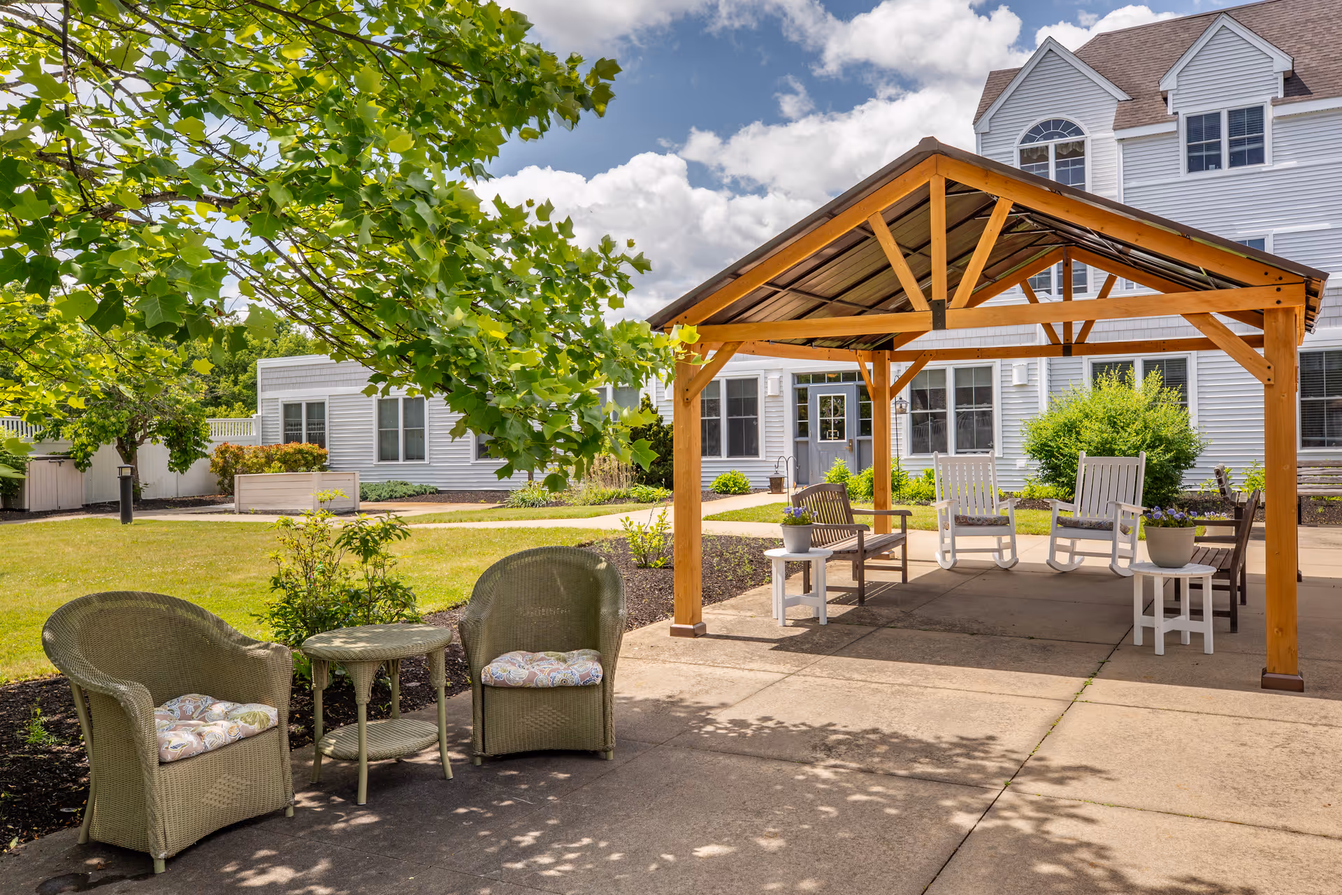 Outdoor seating area at Brookdale Spruce Wood featuring a wooden pergola with rocking chairs and small tables underneath, surrounded by greenery and a white multi-story building in the background under a partly cloudy sky.