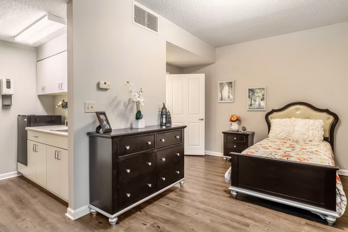 A cozy bedroom in a senior living facility featuring a single bed with a floral bedspread and a cushioned headboard. Next to the bed is a small dark wood nightstand with a vase of flowers and a clock. A matching dark wood dresser with decorative items and a framed photo sits against the wall. The room has light-colored walls, wood flooring, and an open door leading to another area. A small kitchenette with white cabinets and a sink is visible to the left.