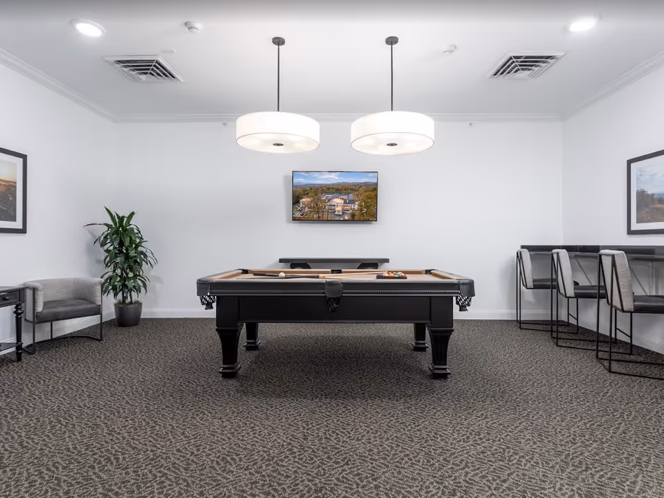 A modern game room with a pool table in the center, two large round pendant lights hanging from the ceiling, a flat-screen TV mounted on the wall displaying an aerial view of a building, a gray armchair and a potted plant on the left, and a high counter with three bar stools on the right. The room has white walls, patterned carpet, and framed pictures on the walls.
