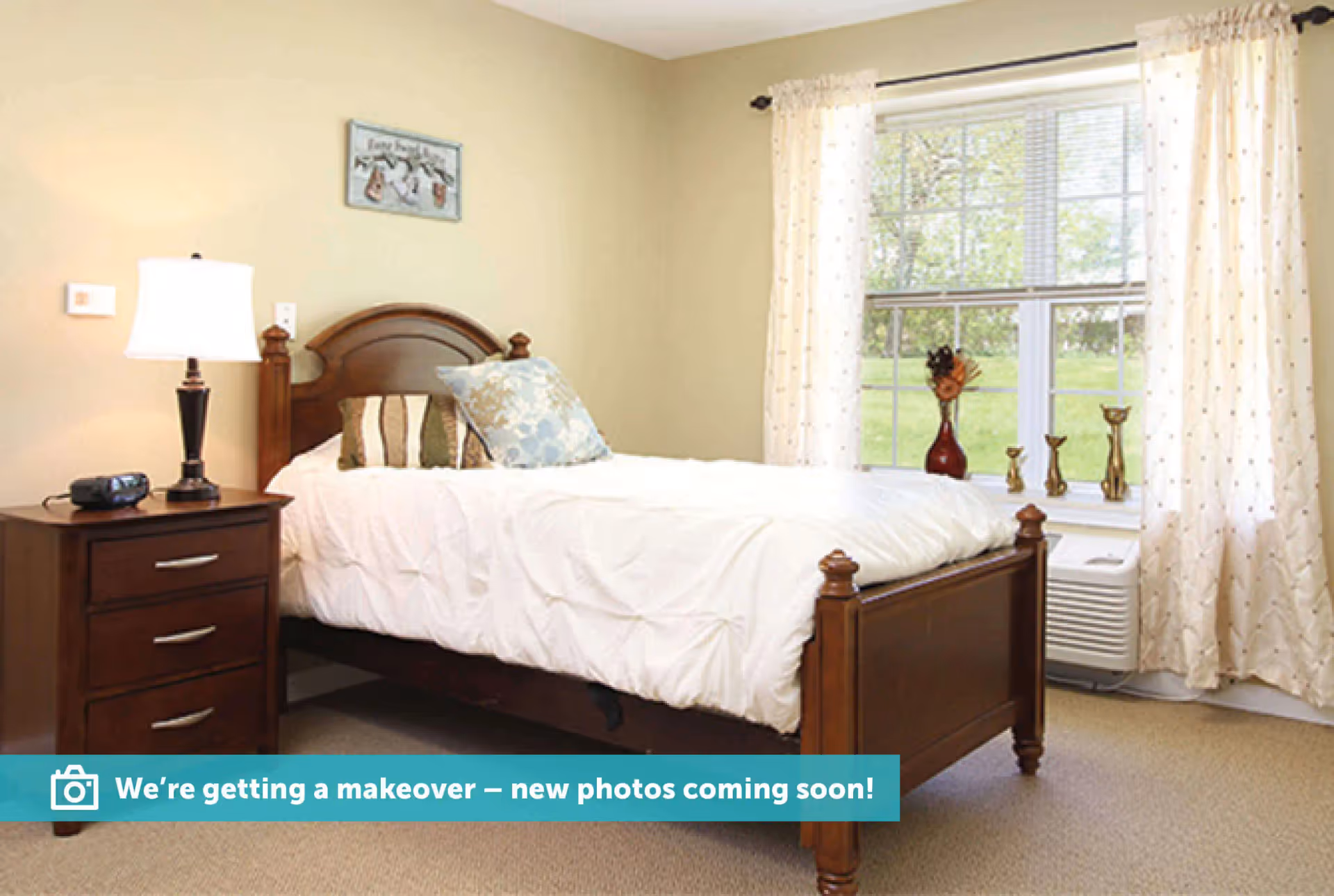 A bedroom with a wooden bed frame and white bedding, two decorative pillows, a wooden nightstand with a lamp and an alarm clock, a window with white curtains, and decorative items on the windowsill.