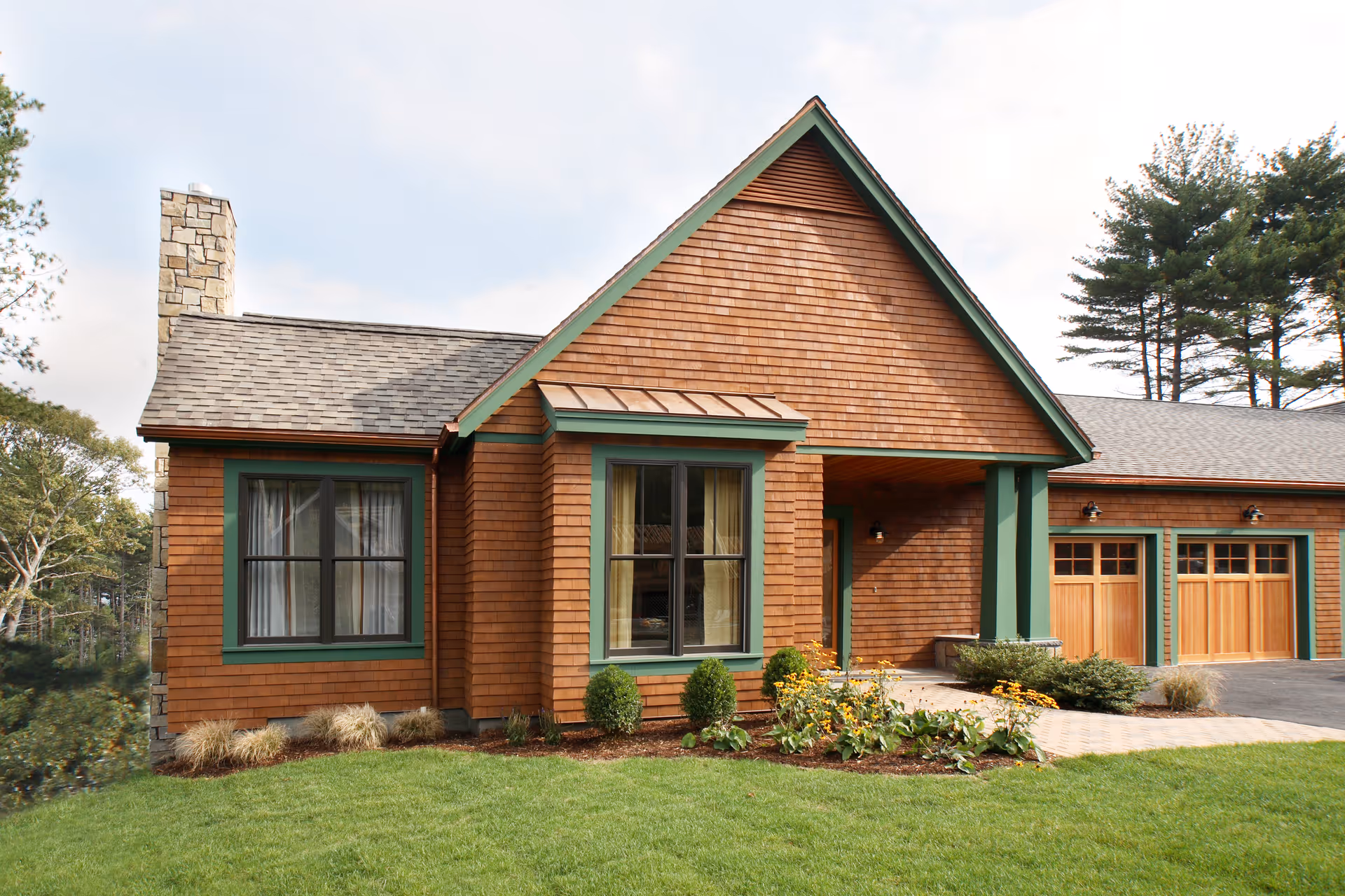 Exterior view of a single-story building with wooden shingles and green trim, featuring a stone chimney, large windows with curtains, a covered entrance, and a three-car garage with wooden doors. The building is surrounded by a well-maintained lawn and some landscaping with bushes and flowers.