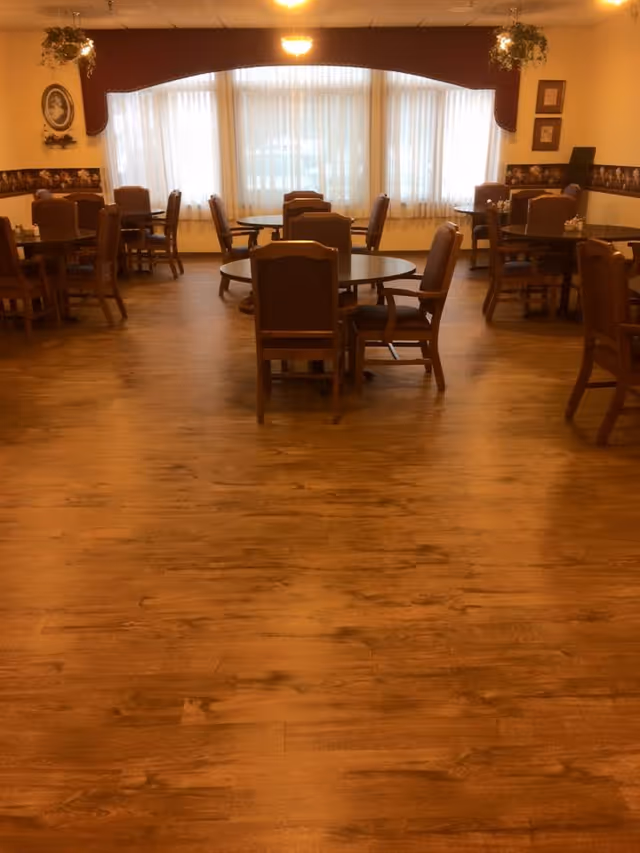 A dining room with several round wooden tables and cushioned chairs arranged around them. The room has wood flooring, large windows with sheer white curtains, and decorative valances. There are framed pictures and hanging plants on the walls.