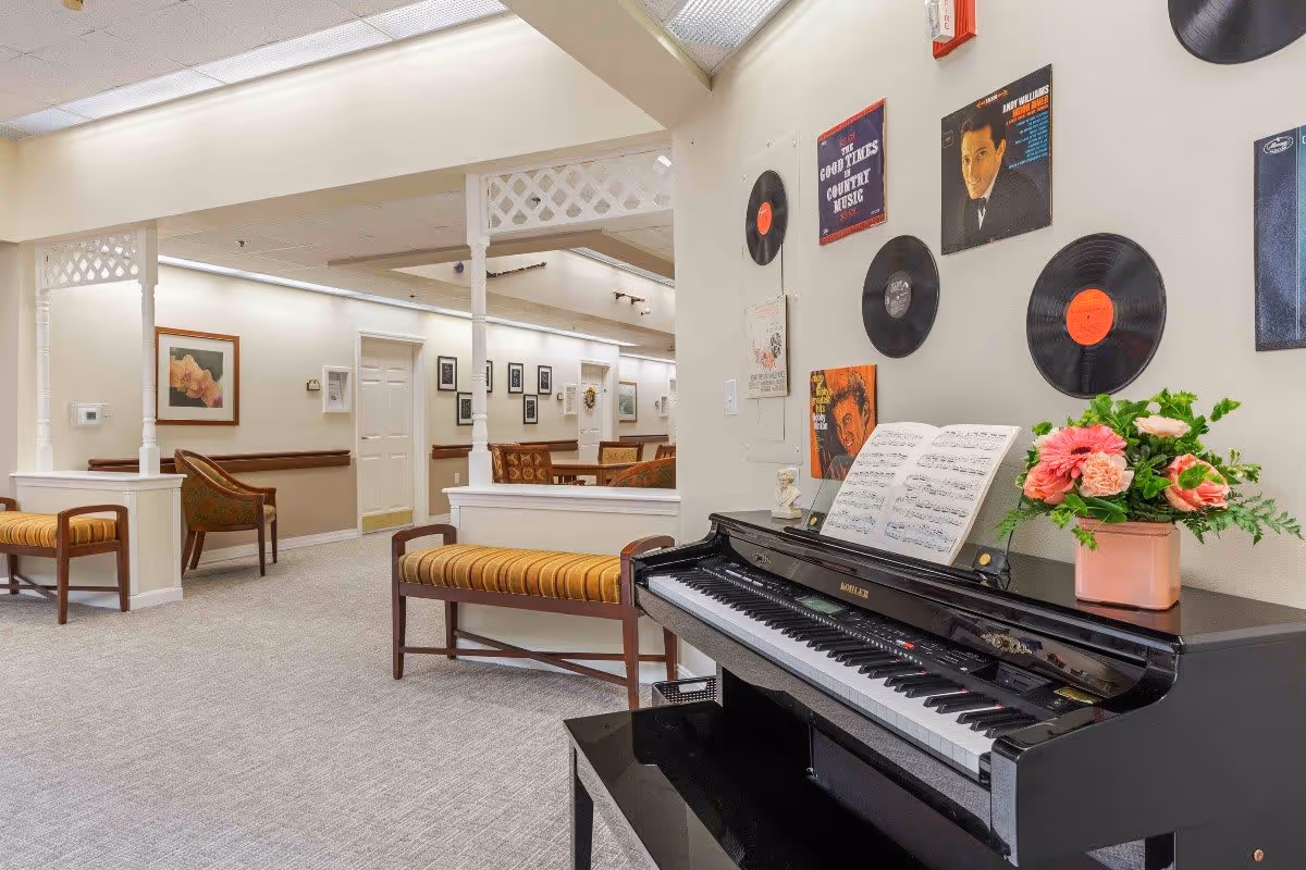 Interior view of a senior living facility lounge area featuring a black piano with sheet music and a flower pot on top. The wall behind the piano is decorated with vinyl records and vintage music album covers. The space includes cushioned benches with striped upholstery and chairs, with framed artwork on the walls and a carpeted floor.