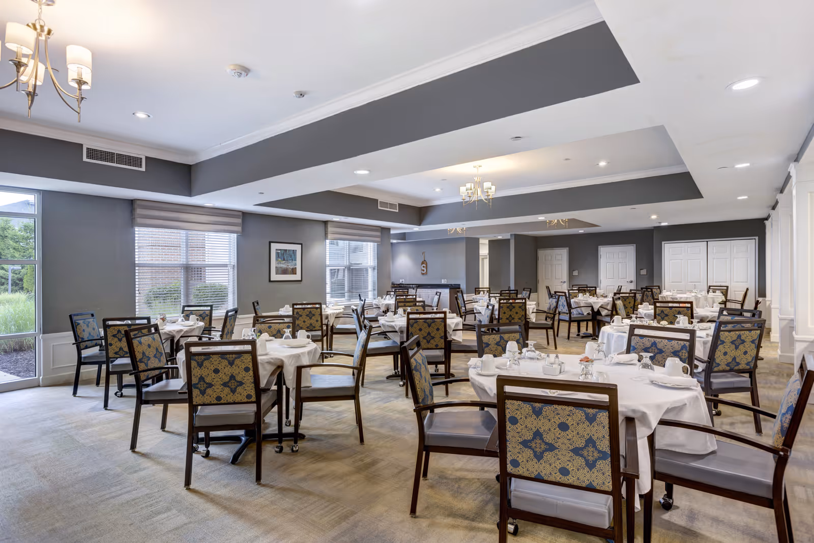 A spacious dining room with multiple round tables covered with white tablecloths and set with glassware and napkins. The chairs have patterned upholstery and wooden frames. Large windows allow natural light to brighten the room, and there are chandeliers hanging from the ceiling. The walls are painted gray with white trim, and the carpet is light-colored.