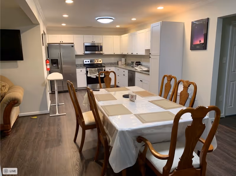 A dining area with a rectangular table covered with a white tablecloth and six wooden chairs around it. The table has beige placemats and a small centerpiece. In the background, there is a kitchen with white cabinets, a stainless steel refrigerator, stove, microwave, and dishwasher. To the left, part of a beige sofa and a wall-mounted TV are visible. The room has wooden flooring and neutral-colored walls.