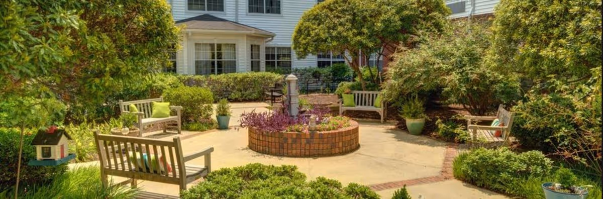 Sunny landscaped courtyard with wooden benches arranged around a circular brick planter in front of a residential building.