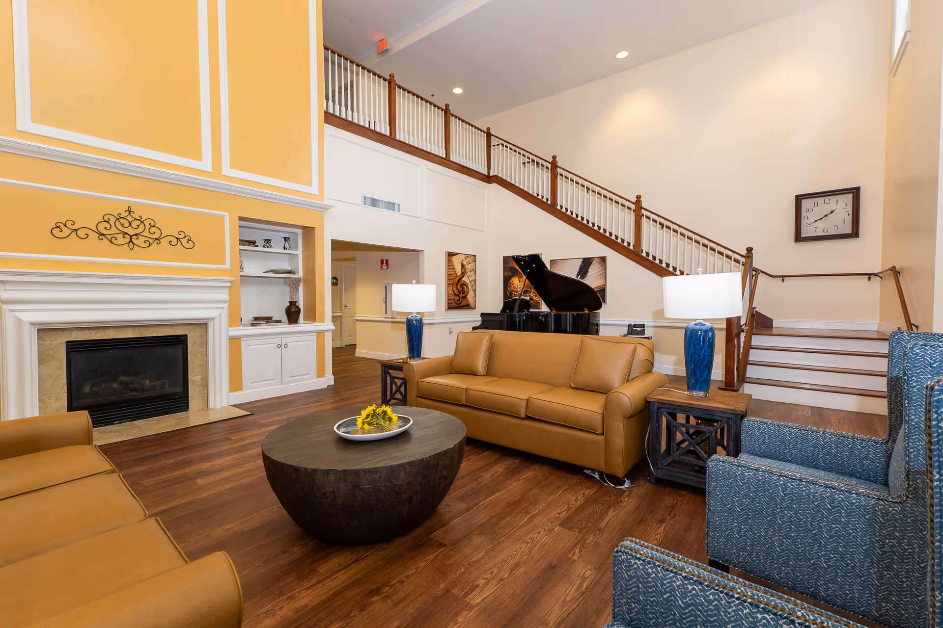 A spacious senior living facility common area with high ceilings, featuring a brown leather sofa, two blue armchairs, a round wooden coffee table with a decorative plate and flowers, a fireplace with a yellow accent wall, built-in white shelves, a grand piano, and a staircase with wooden handrails.