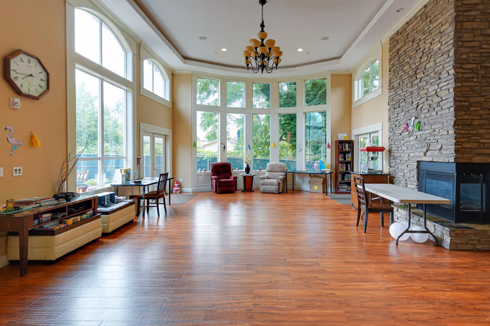 A spacious senior living common area with large floor-to-ceiling windows letting in natural light. The room features wooden flooring, a stone fireplace on the right, two recliner chairs near the windows, tables and chairs along the walls, a clock on the left wall, and a chandelier hanging from the ceiling.