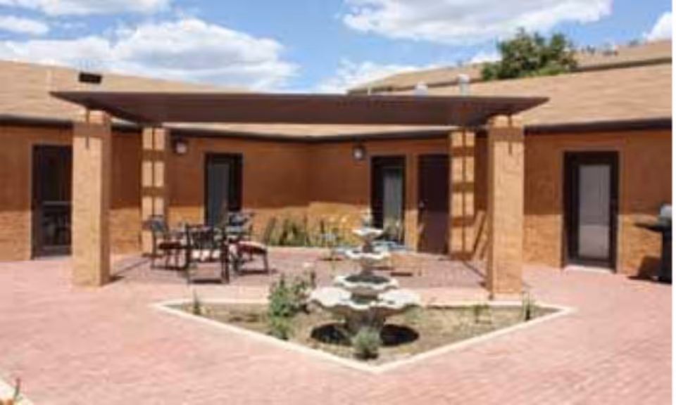 Outdoor courtyard area with a covered pergola, patio chairs and tables, a multi-tiered water fountain in the center, surrounded by a brick-paved ground and brown building walls with doors.