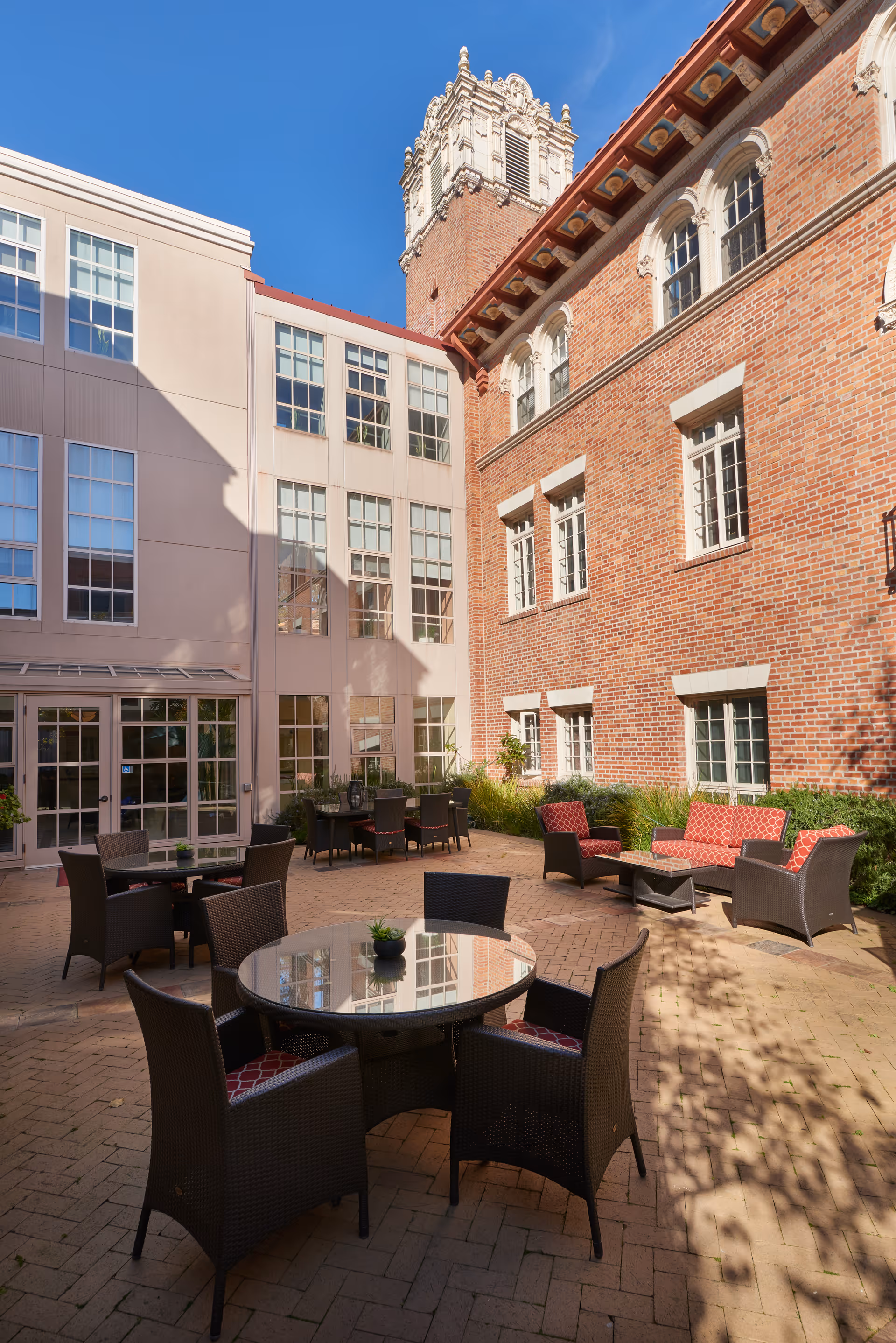 Outdoor courtyard area with several round glass-top tables and wicker chairs with red cushions. There are also wicker sofas and armchairs with red cushions arranged around a coffee table. The courtyard is surrounded by a brick building on one side and a beige building with many windows on the other side. The sky is clear and blue.
