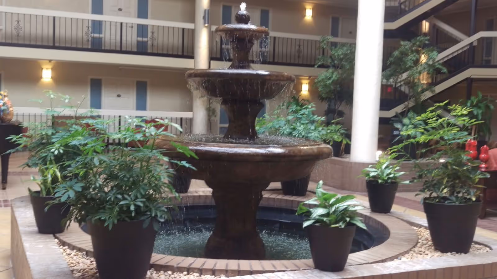 Indoor three-tiered water fountain surrounded by potted green plants in a spacious atrium with railings and doors on the upper floors visible in the background.