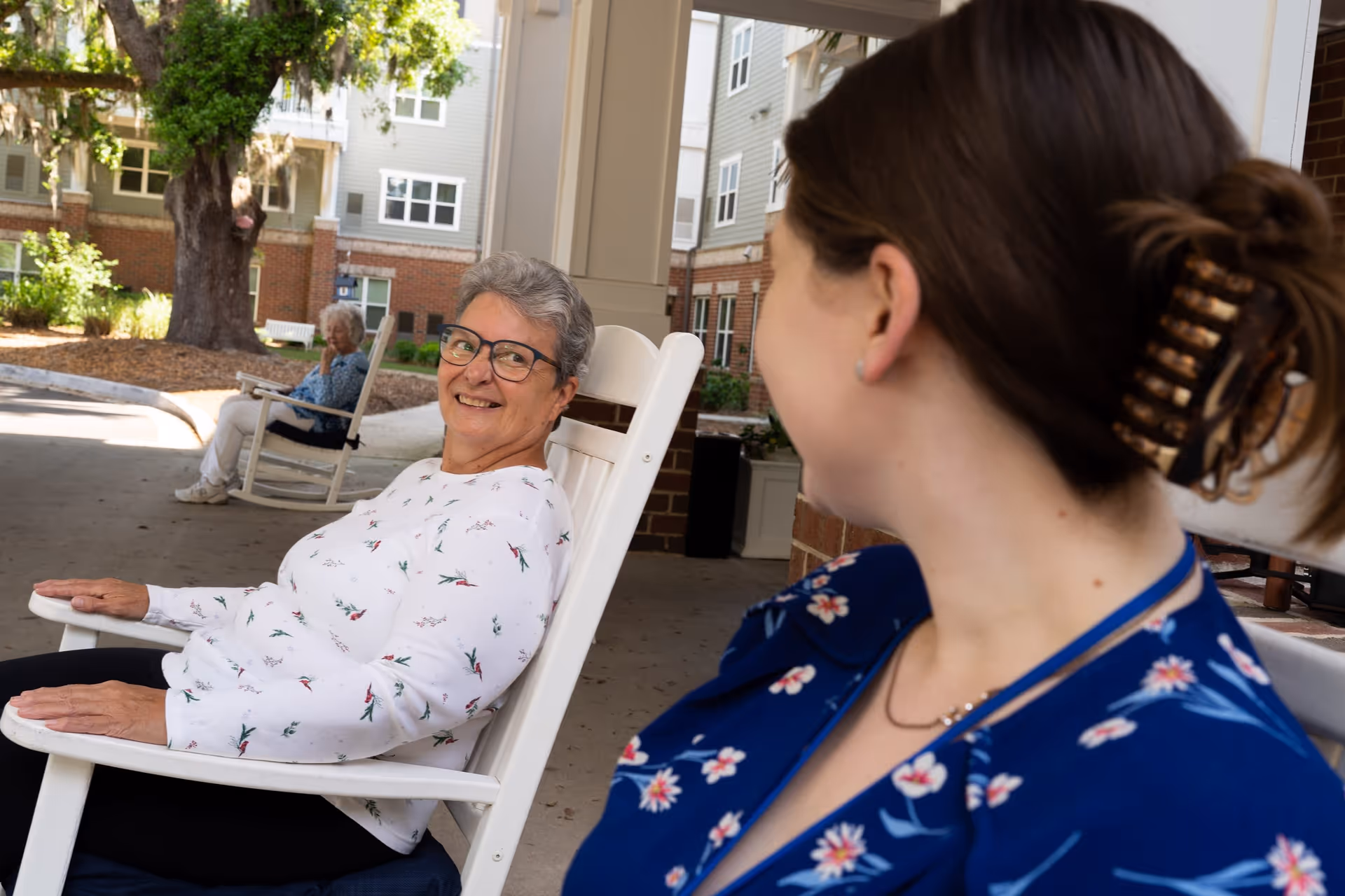 Two women sitting and smiling at each other on white rocking chairs outside a senior living facility. One woman has short gray hair and glasses, wearing a white floral top, and the other has brown hair tied back with a hair clip, wearing a blue floral top. In the background, another elderly woman is sitting on a rocking chair near a large tree and the building.