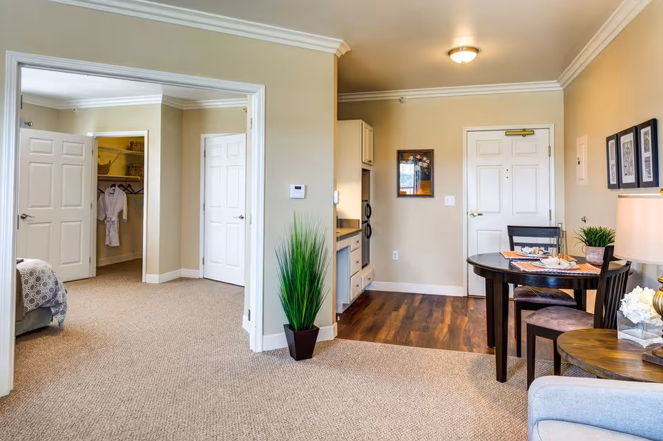 Interior view of a senior living facility apartment showing a small dining area with a round table and two chairs, a kitchenette with cabinets and appliances, and an open doorway leading to a bedroom with a walk-in closet. The room has beige walls, carpeted and wooden floors, and decorative plants and framed pictures on the walls.