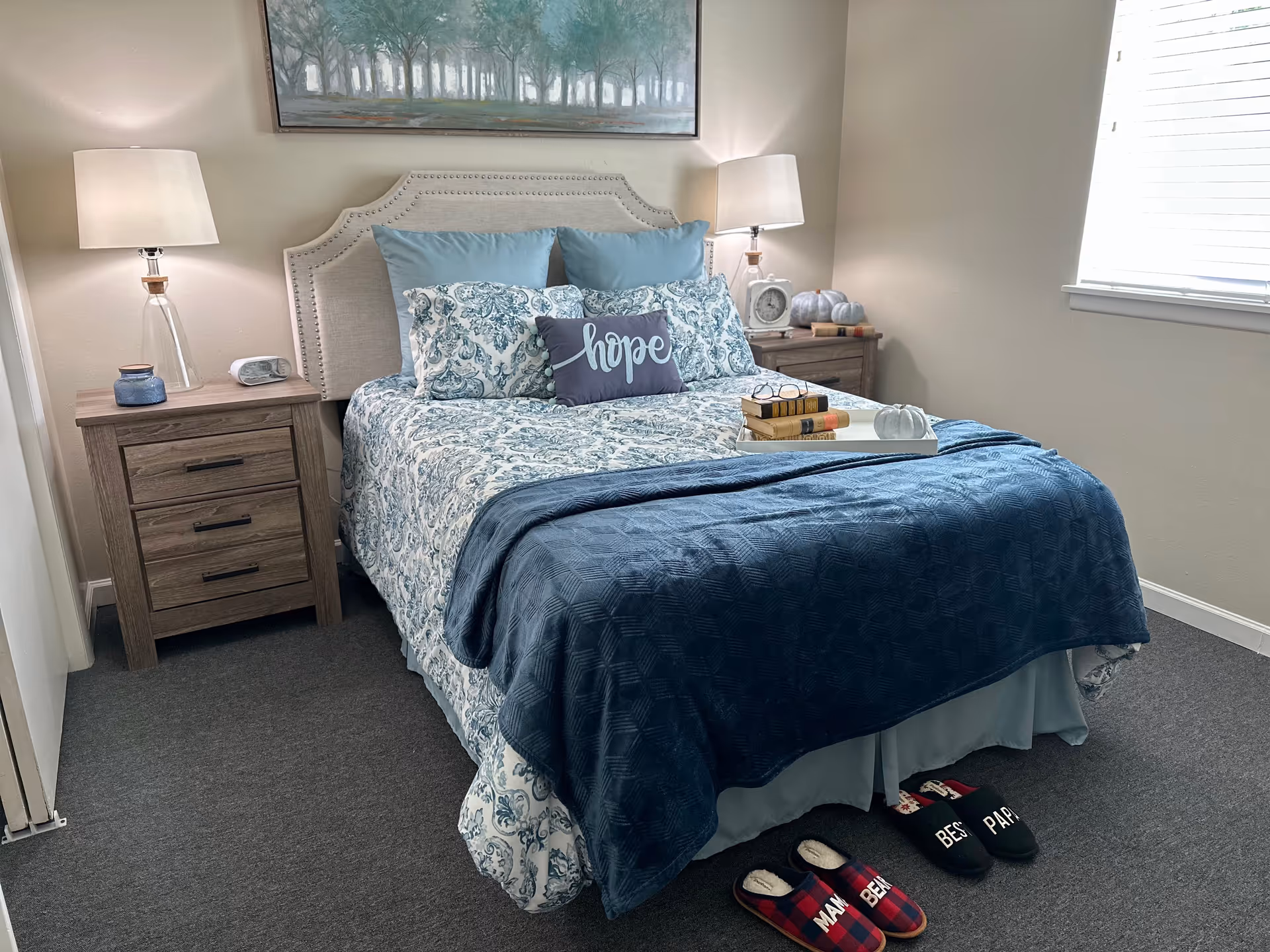 Neatly made bed in a softly lit bedroom with blue patterned bedding, matching nightstands and lamps, artwork above, and slippers on the floor.