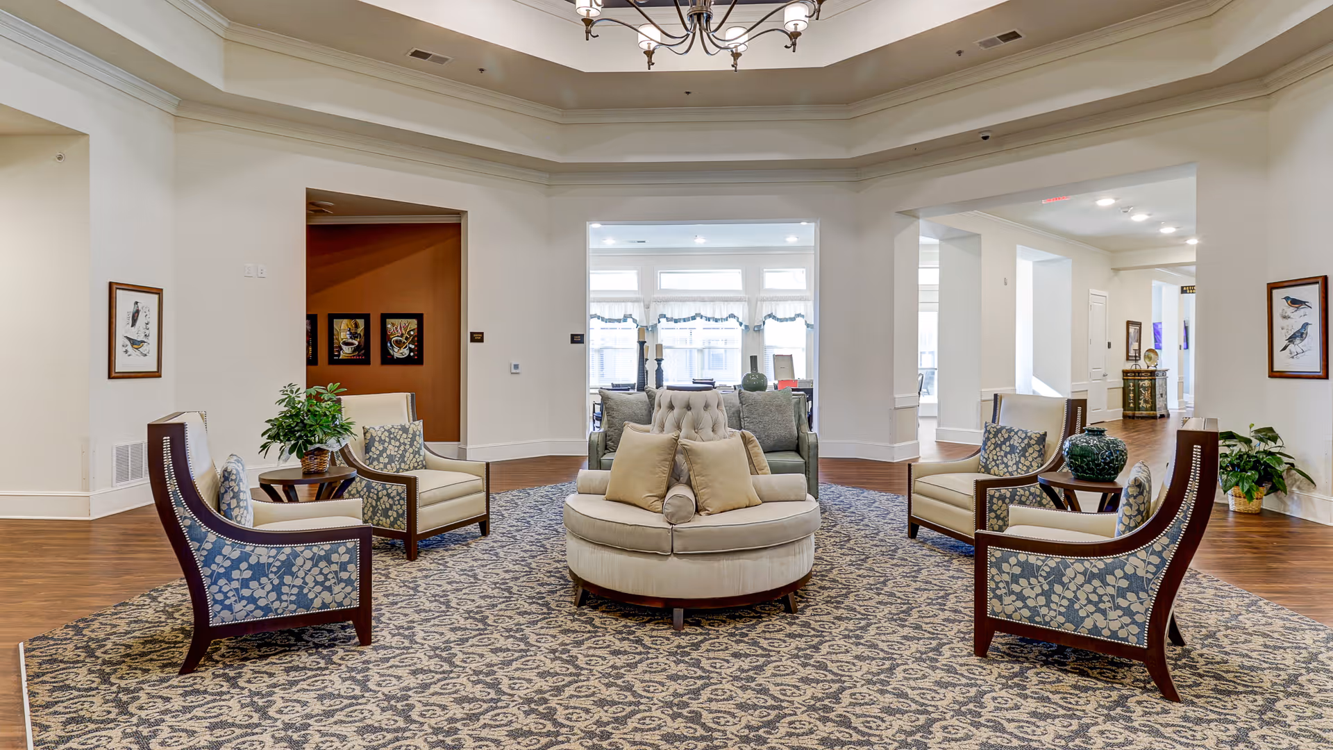 A spacious and elegant senior living facility common area with a circular beige cushioned seating arrangement in the center, surrounded by four armchairs with floral upholstery. The room features a patterned carpet, hardwood floors, white walls, framed bird artwork, potted plants, and a chandelier hanging from a tray ceiling. Large windows in the background allow natural light to fill the space.