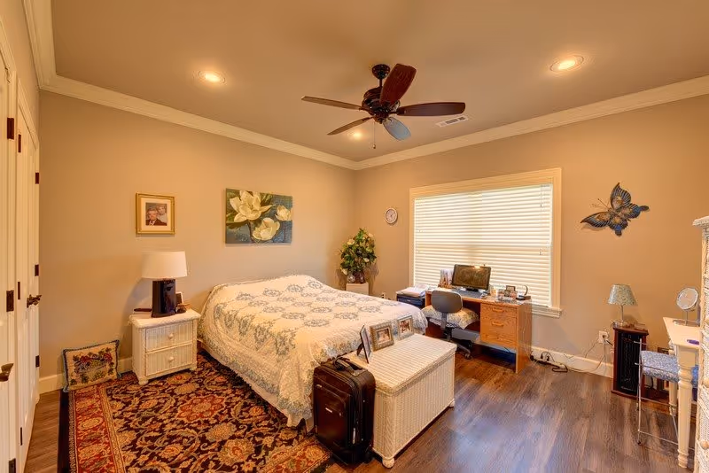 A cozy bedroom with a large bed covered in a white and blue patterned quilt. The room features a ceiling fan with lights, a wooden floor with a decorative rug, a wicker nightstand with a lamp, and a wicker chest at the foot of the bed. There is a desk with a computer and chair near a window with closed blinds. The walls are decorated with framed pictures and a butterfly wall ornament. A potted plant is placed in the corner.