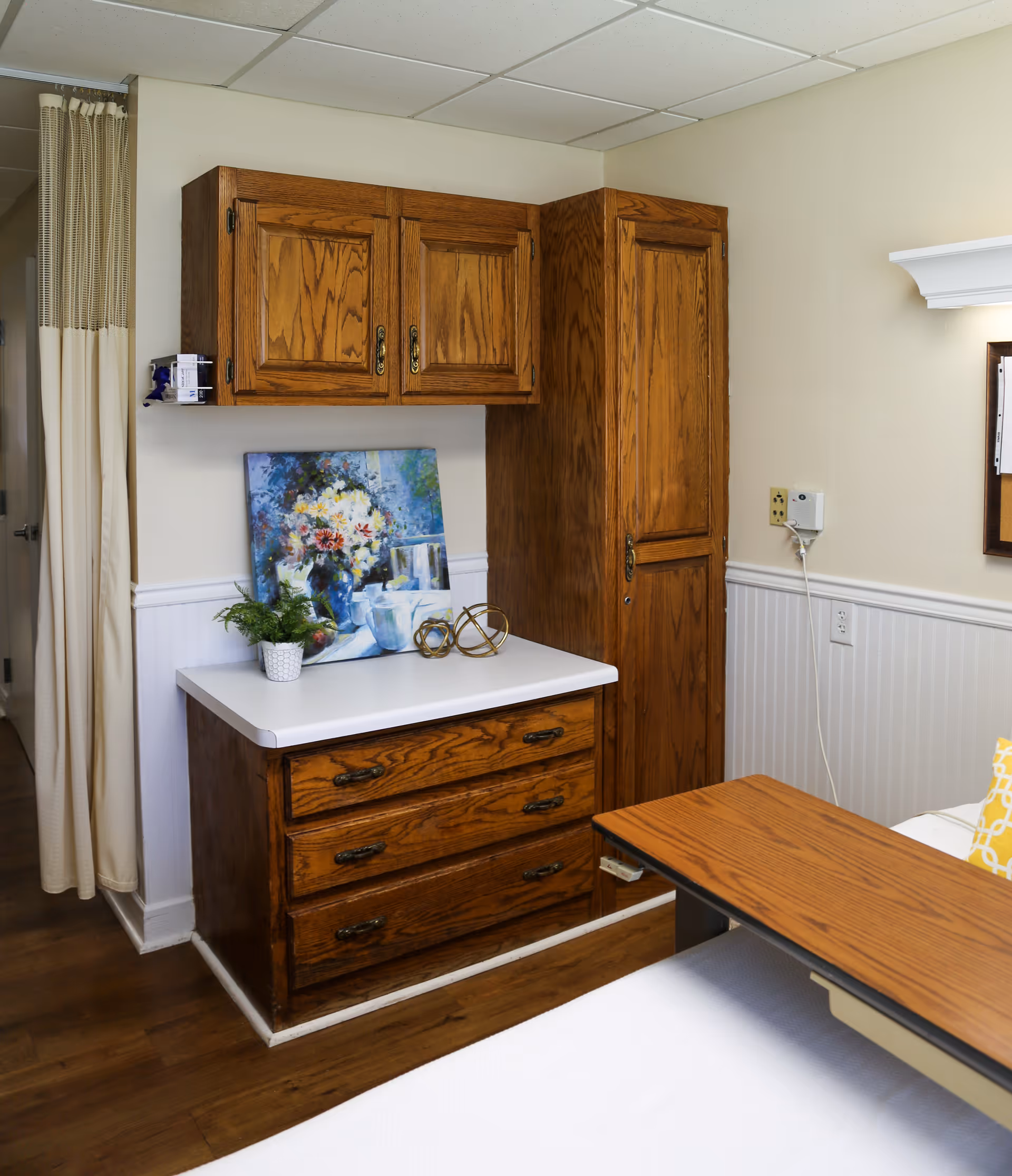 A bedside area in a senior room with wooden cabinets and drawers, a small countertop with decorations, a retractable overbed table, and a privacy curtain.