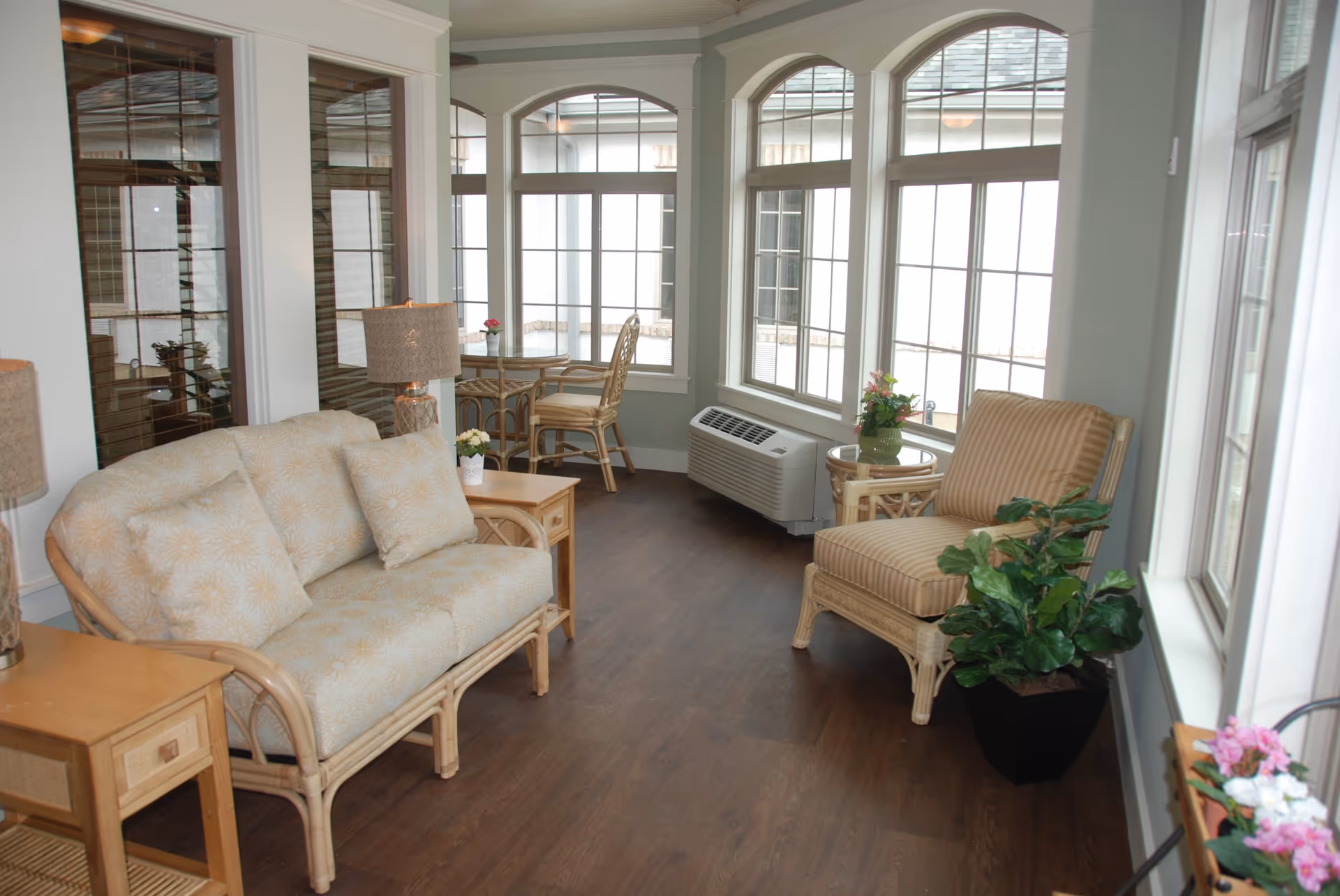 Bright sunroom-style sitting area with wicker sofa and chairs, a small table, plants, and large arched windows.