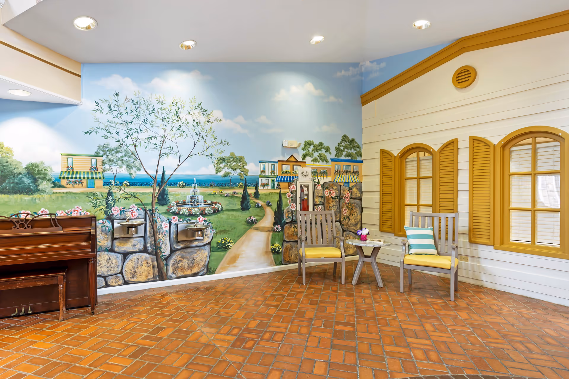 Indoor seating area with two wooden chairs with yellow cushions and a small round table with a flower pot. The wall behind features a mural depicting a garden path, trees, houses, and a fountain. To the left, there is a brown upright piano. The floor is covered with reddish-brown tiles.